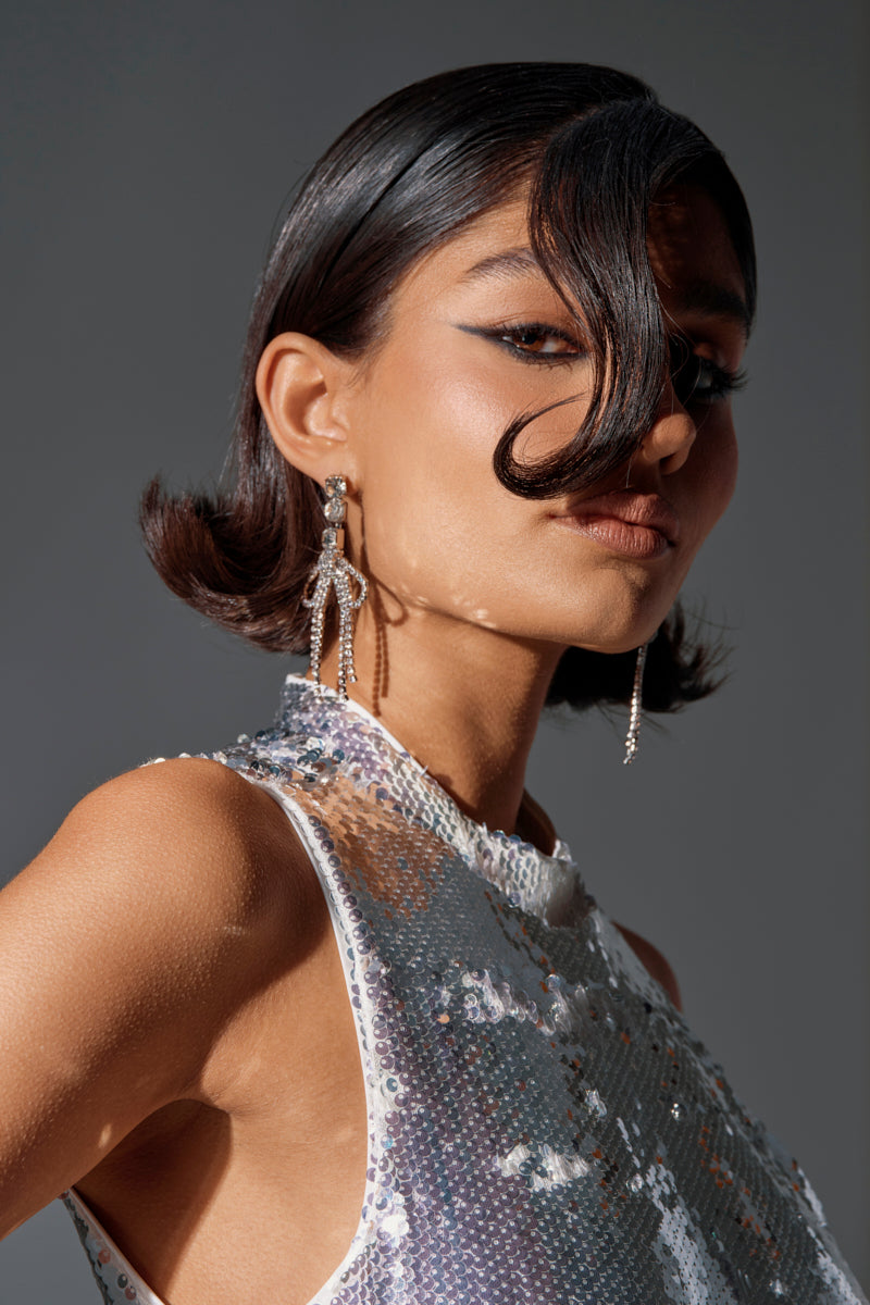 A woman with short, sleek dark hair poses confidently wearing the WITH A BOW SILVER EARRING and a shiny silver sequin top. A lock of hair partly covers her face, set against a plain gray background for a chic, modern look.