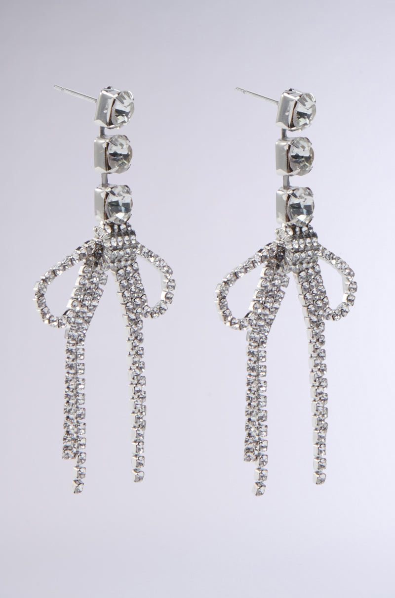 The WITH A BOW SILVER EARRING features three square stones at the top and a dangling bow accent with sparkling rhinestone fringe, adding elegant flair.