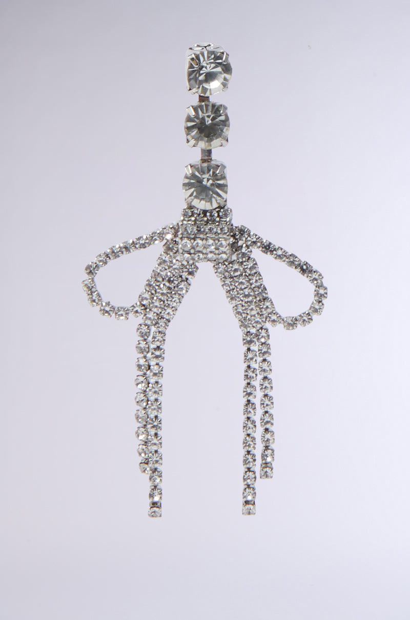 The WITH A BOW SILVER EARRING features a row of round gems at the top and bow attachments with cascading rhinestone fringe, set against a plain light background.