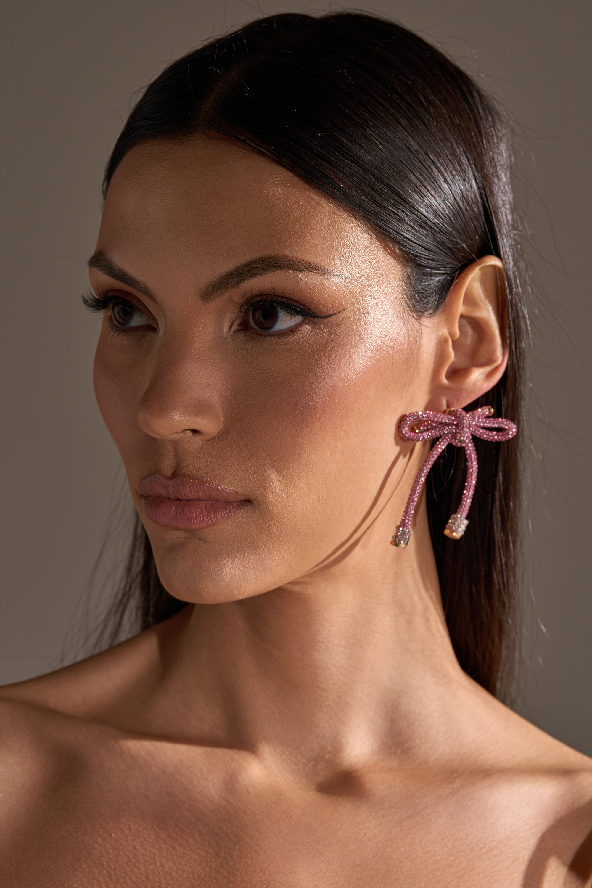 A woman with straight dark hair wears the ALL NIGHT LONG LIGHT PINK EARRING—large, sparkling pink rhinestone bow earrings. She has smooth skin, subtle makeup, a neutral expression, and is set against a softly lit plain background. 2