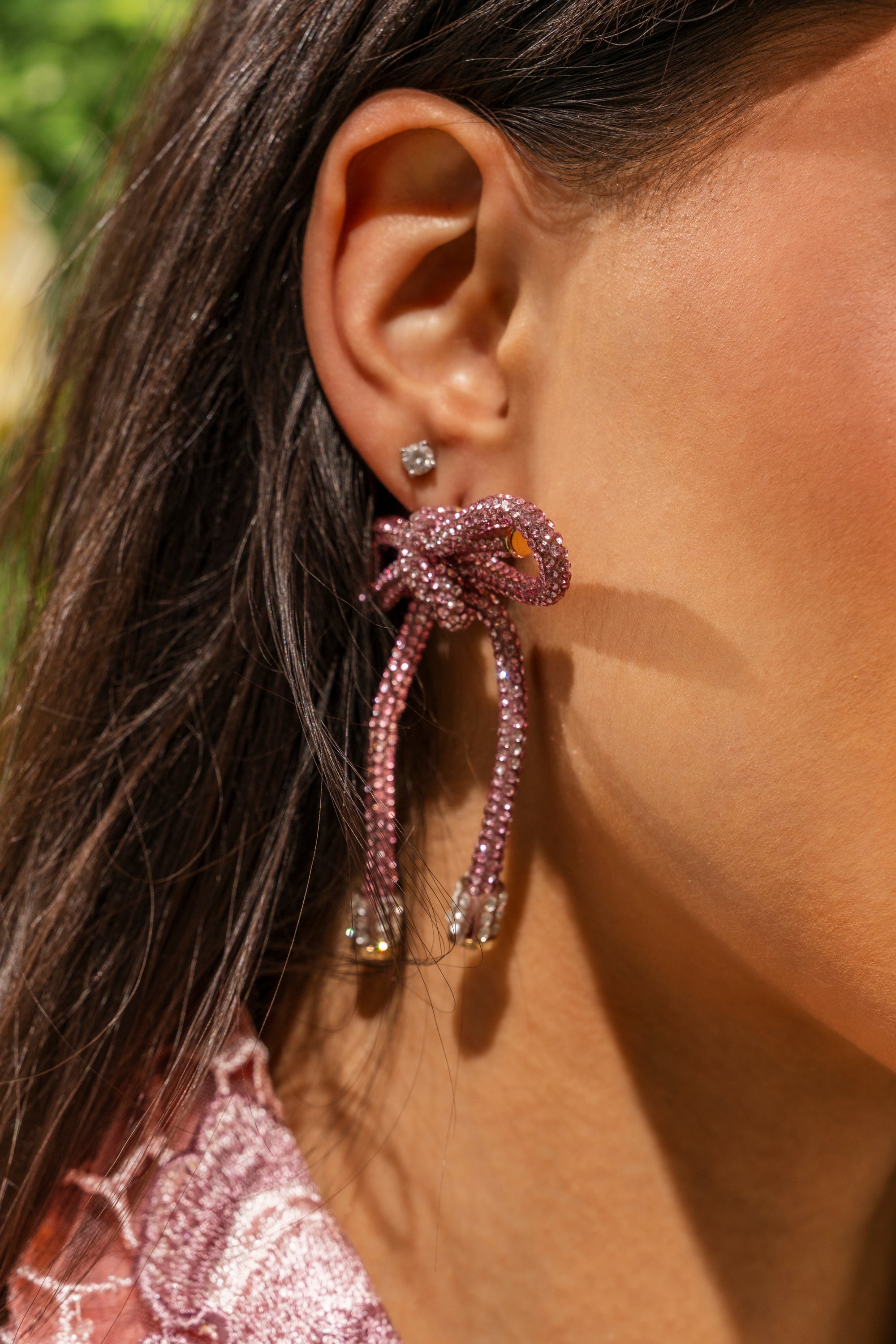 Close-up of a woman's ear with dark hair tucked behind it, wearing two earrings: a small square stud and the ALL NIGHT LONG LIGHT PINK EARRING—an oversized pink rhinestone bow earring shaped like a knot with dangling strands. 0