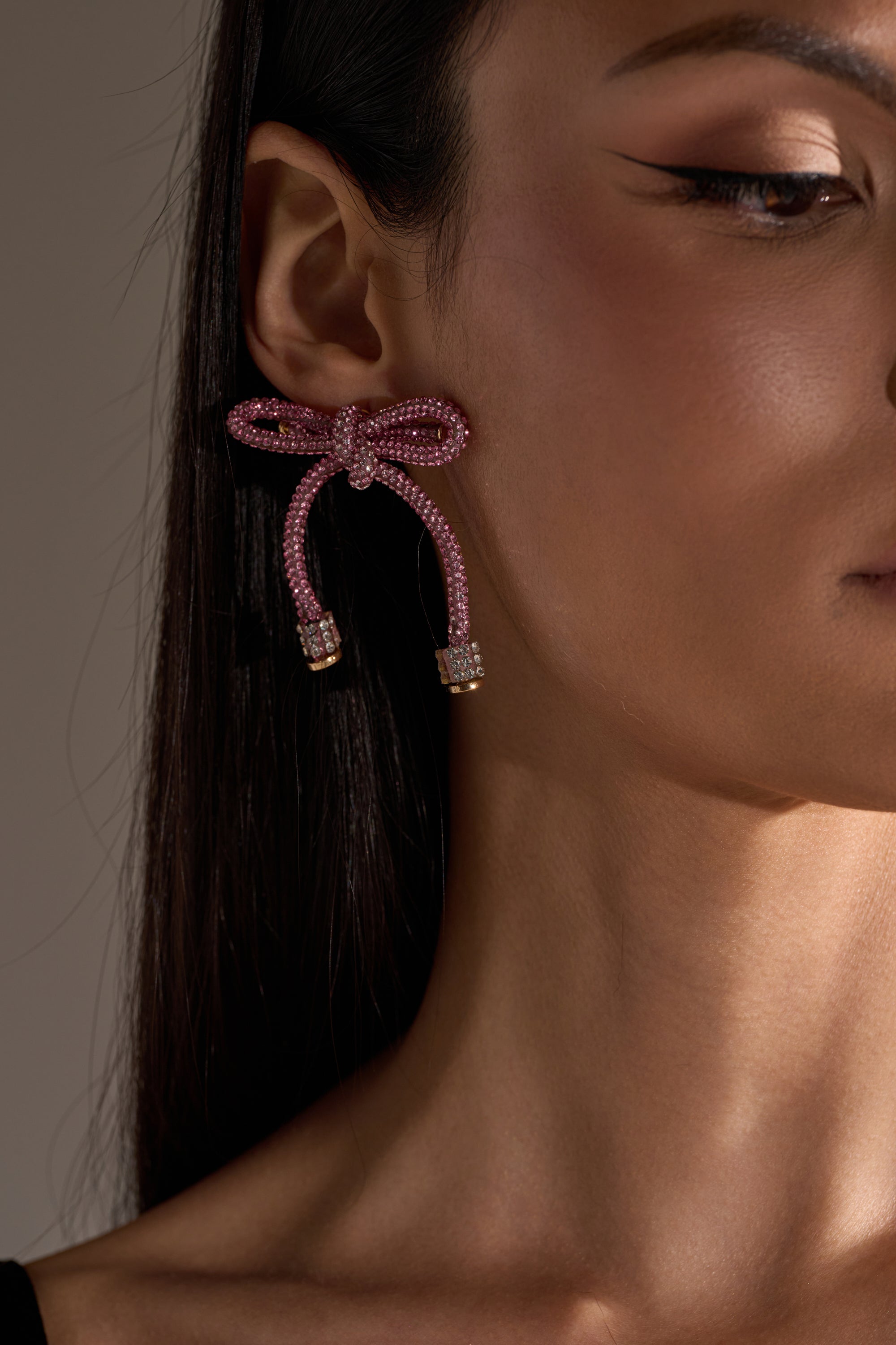 A close-up of a woman wearing the ALL NIGHT LONG LIGHT PINK EARRING. Her straight dark hair is tucked behind her ear, showing off the large pink bow design. She has winged eyeliner and smooth, glowing skin. 3