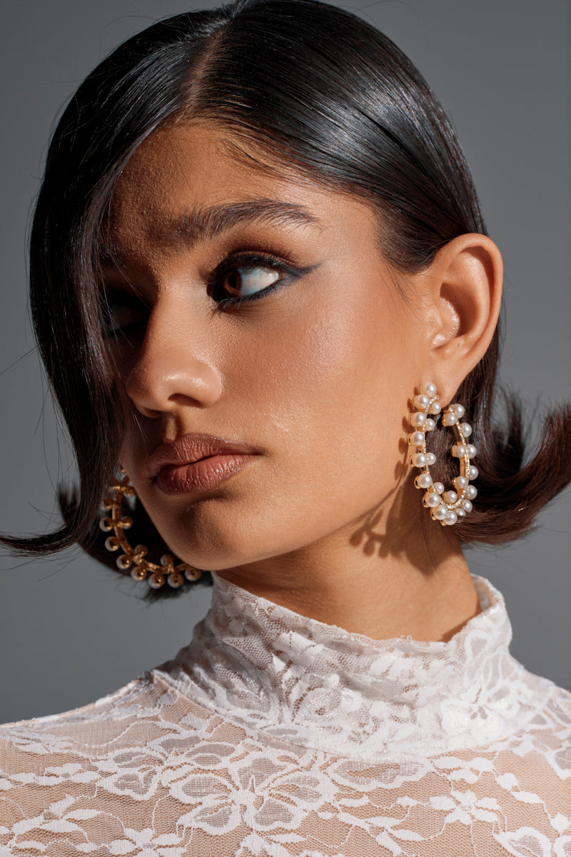 A woman with smooth, dark bobbed hair and bold black eyeliner wears the EVERYTHING YOU WANT EARRING—large hoops with subtle pearl detail—and a white lace high-neck top, gazing to the side with a neutral expression. 0