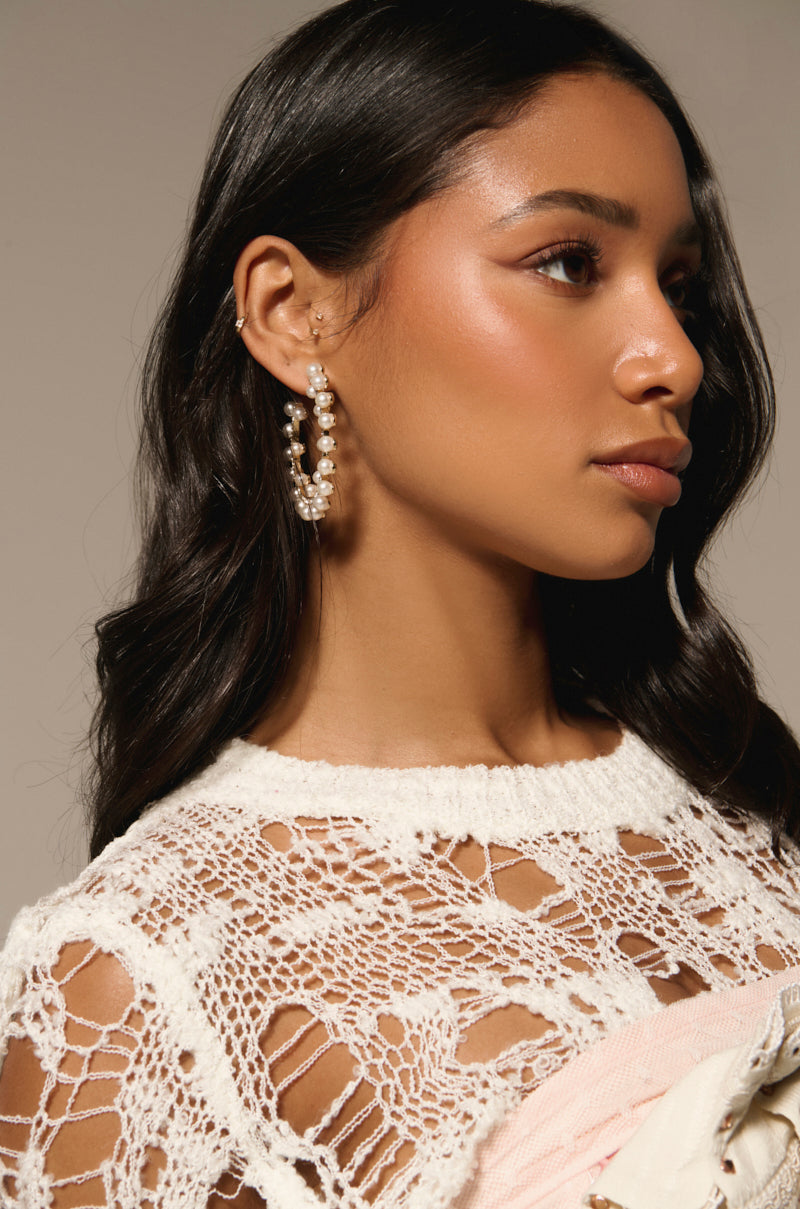 A woman with long dark hair wearing a white crocheted top and pearl hoop earrings by EVERYTHING YOU WANT EARRING gazes to the side with a neutral expression. The background is soft beige. 2