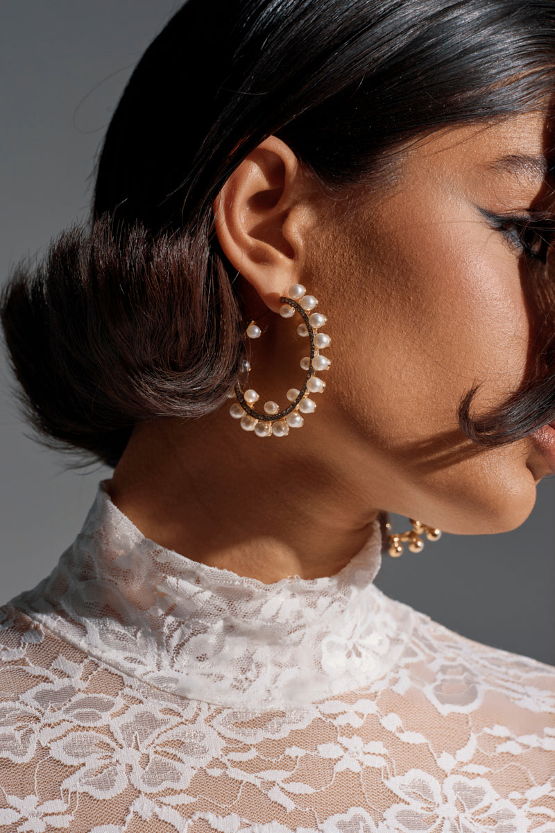 Close-up of a woman in a sheer white lace top wearing the EVERYTHING YOU WANT EARRING—large faux gold hoops with pearl details. Her dark wavy hair and smooth skin are accentuated by elegant lighting. 1