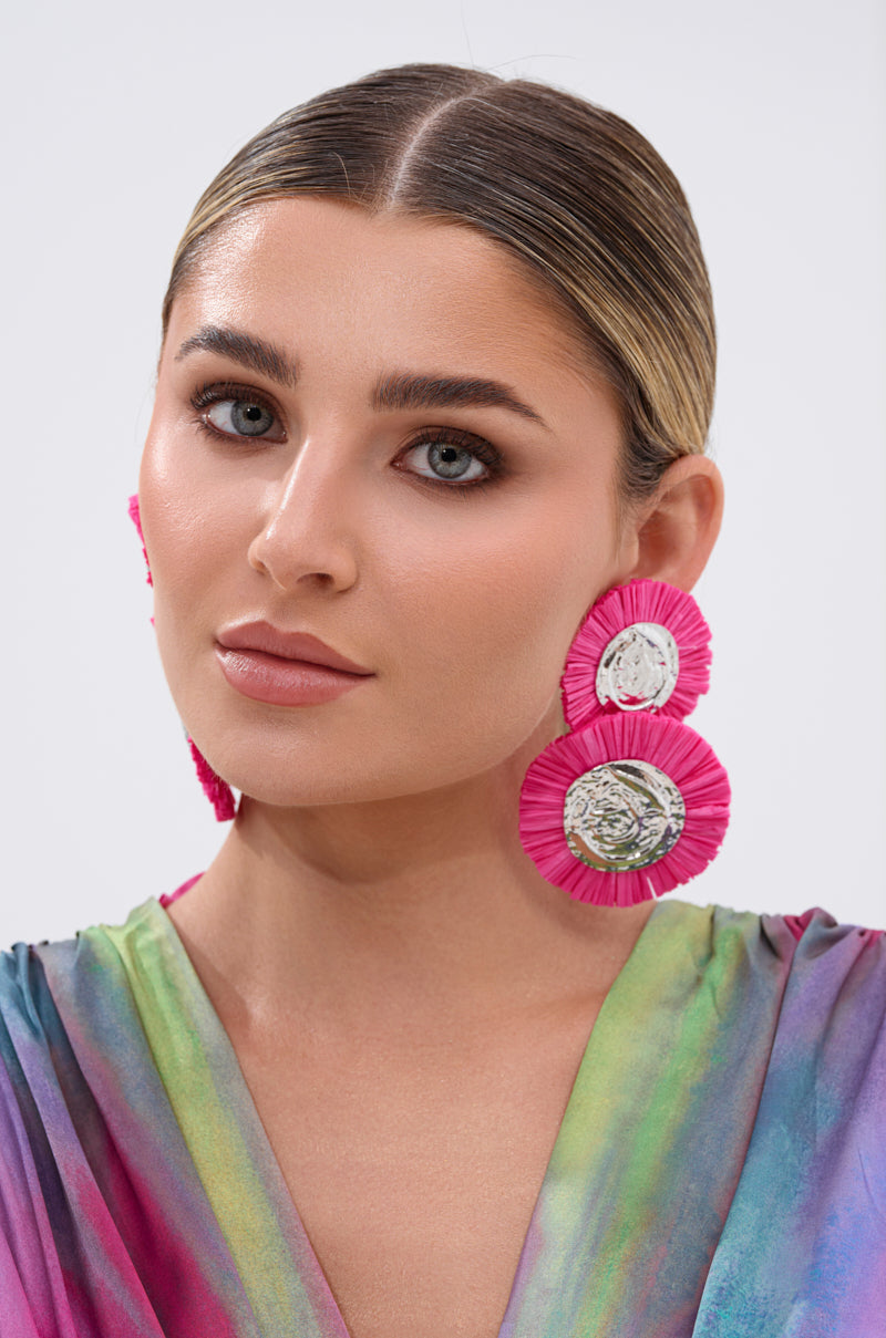 A woman with slicked-back hair wears the IN MIAMI EARRING—hot pink raffia statement earrings—and a colorful, iridescent top, posing against a plain light background. 1
