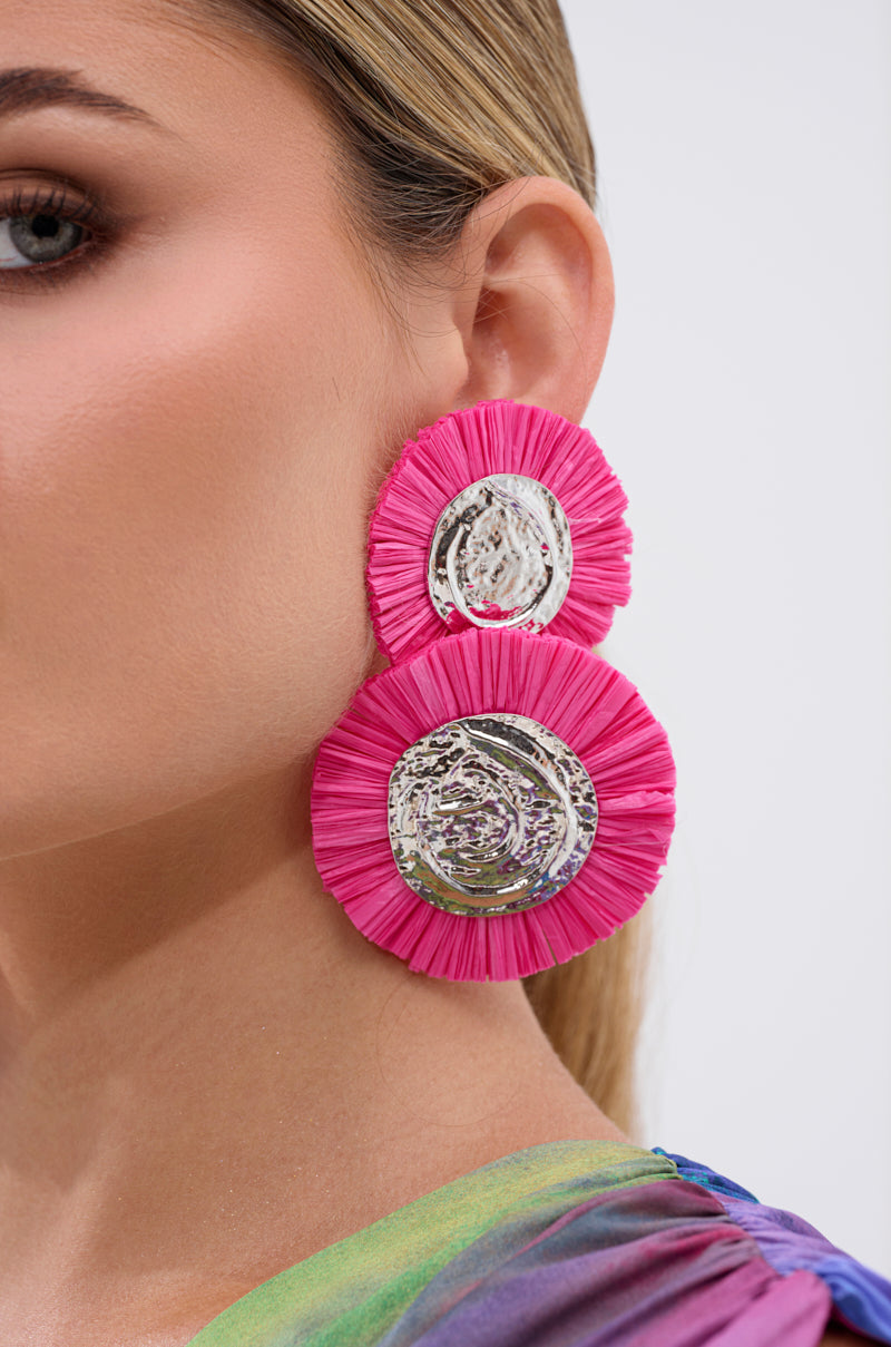 Close-up of a woman's ear wearing the IN MIAMI EARRING, featuring large bright pink fringes and shiny silver circles. Her hair is pulled back, and she has on a colorful iridescent top. 2