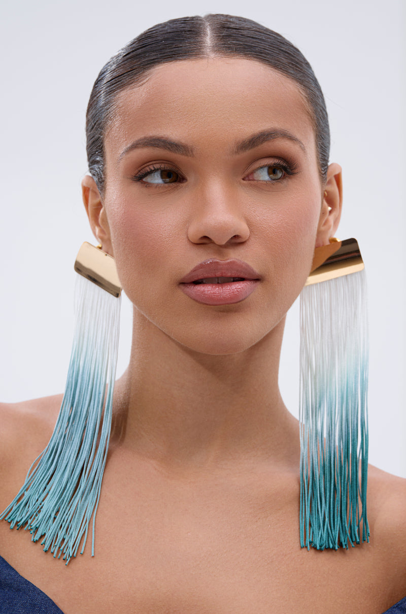 A woman with smooth, pulled-back hair wears large REBRAND EARRING statement earrings featuring gold tops and ombre tassel fringe fading from white to turquoise. She has natural makeup and glances slightly to the side.