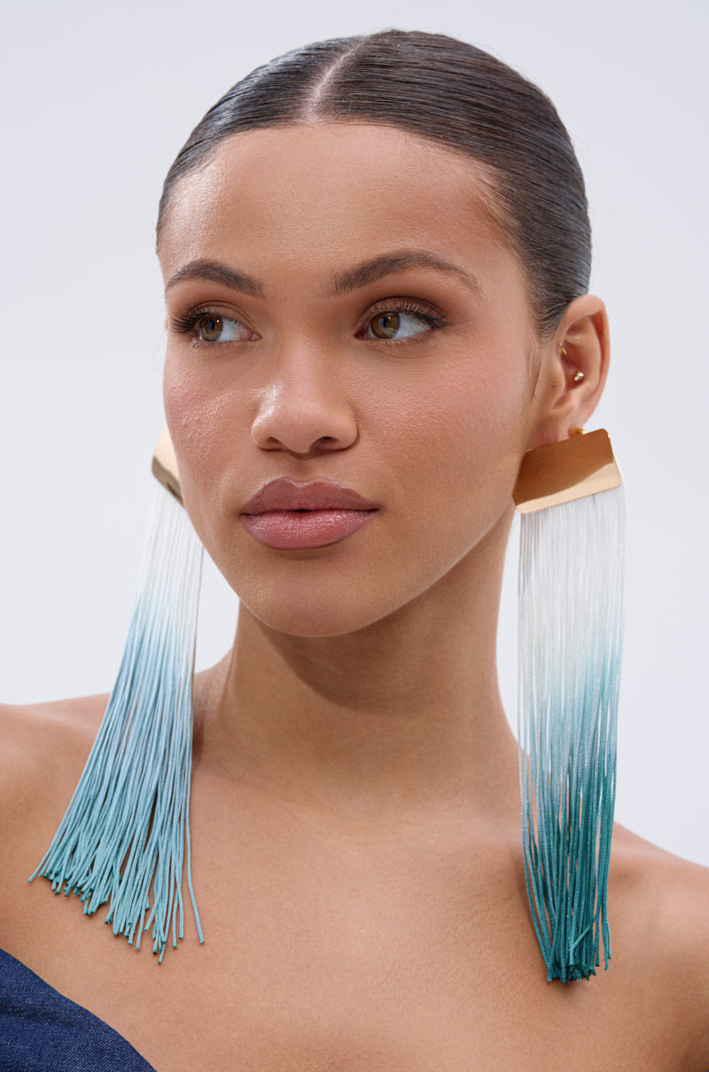 A woman with smooth, pulled-back hair wears REBRAND EARRING statement earrings with long gradient blue-and-white tassel fringe and gold geometric tops, looking slightly to the side against a plain background. 2