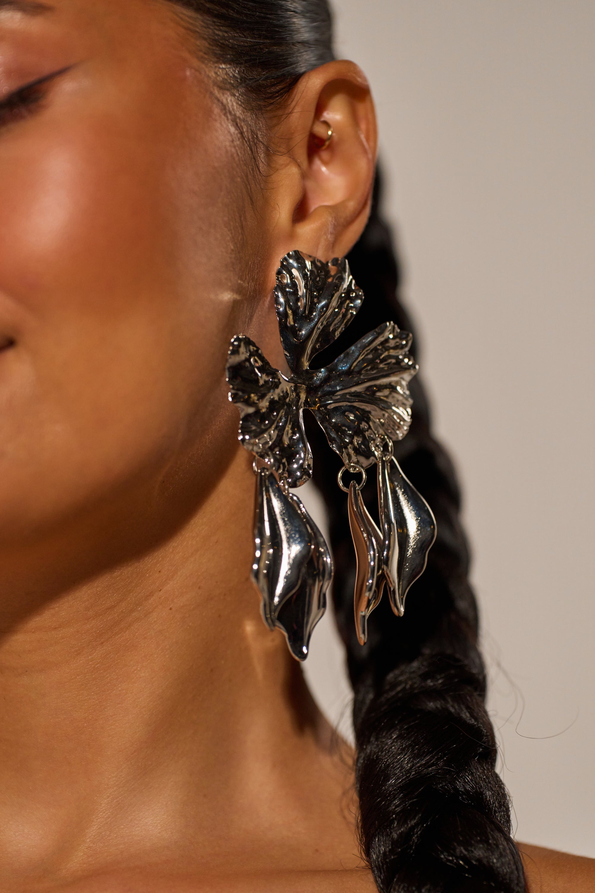Close-up of a woman with a braid wearing the IN BLOOM EARRING—oversized, silver-tone floral statement earrings in an abstract leaf shape that catch the light and complement her glowing skin. 3
