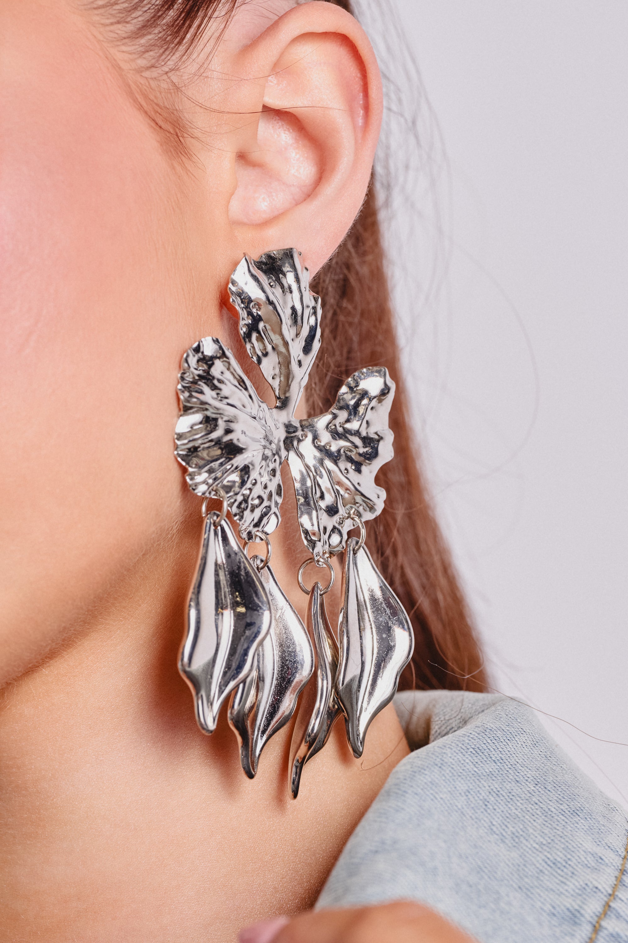 Close-up of a person wearing the IN BLOOM EARRING, a large silver-tone piece shaped like a butterfly with flowing, leaf-like details. The striking earring contrasts against their ear, cheek, and denim collar. 1