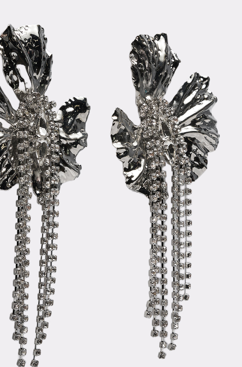 The ON A TRIP EARRING is a shiny silver-tone drop earring shaped like an abstract flower, with rhinestone tassels and sparkling crystal strands, shown against a simple light background. 3