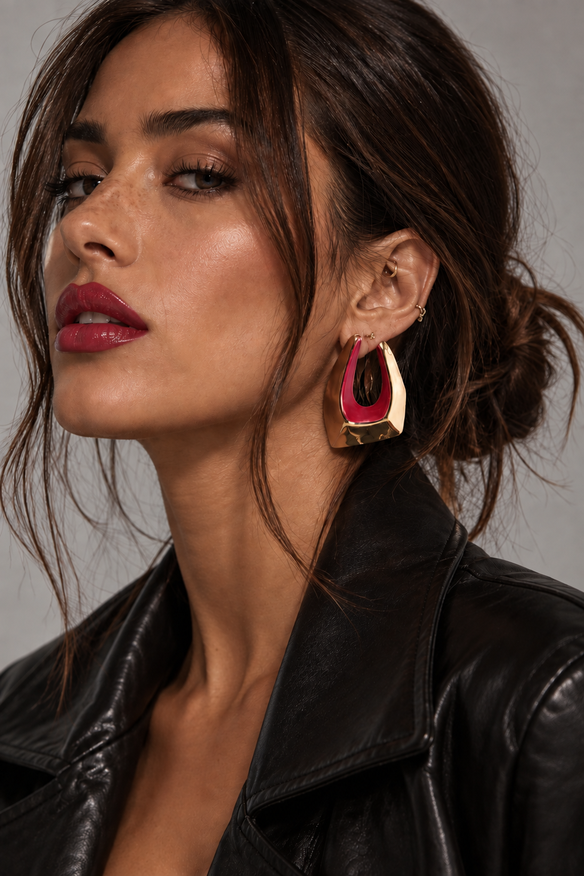 A woman wearing RED FLAG ENERGY EARRINGS, which are sculptural, gold-tone drop earrings featuring a large U-shaped hoop with a pink interior.