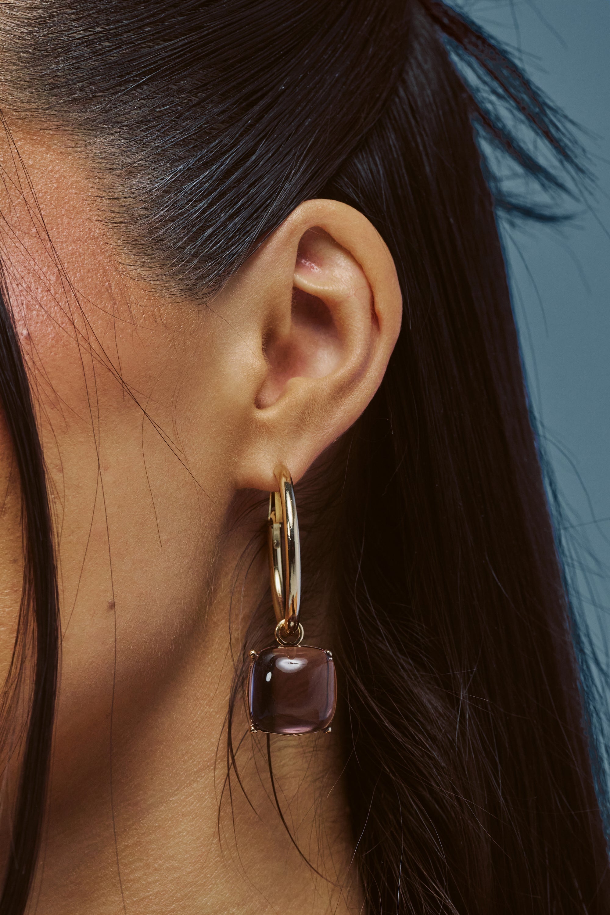 Close-up of a person’s ear with long dark hair, wearing the PURPLE HAZE DROP HOOPS: large gold hoop earrings adorned with a square purple gemstone pendant, set against a blue background.