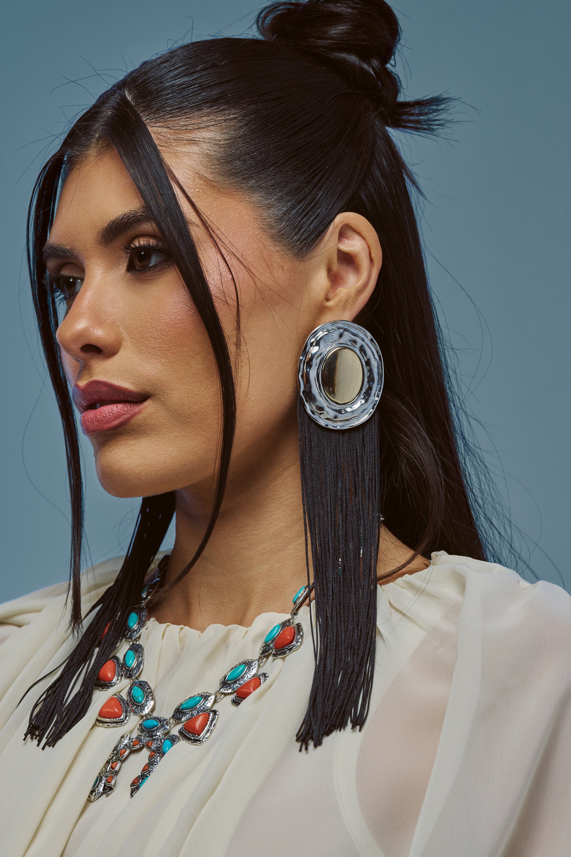 A woman with long dark hair styled half-up, wearing a cream blouse and the TASSEL DIVA STATEMENT EARRINGS IN BLACK—large circular silver earrings accented with black tassel fringe—along with a necklace featuring turquoise and coral-colored stones, gazes to the side against a blue background.