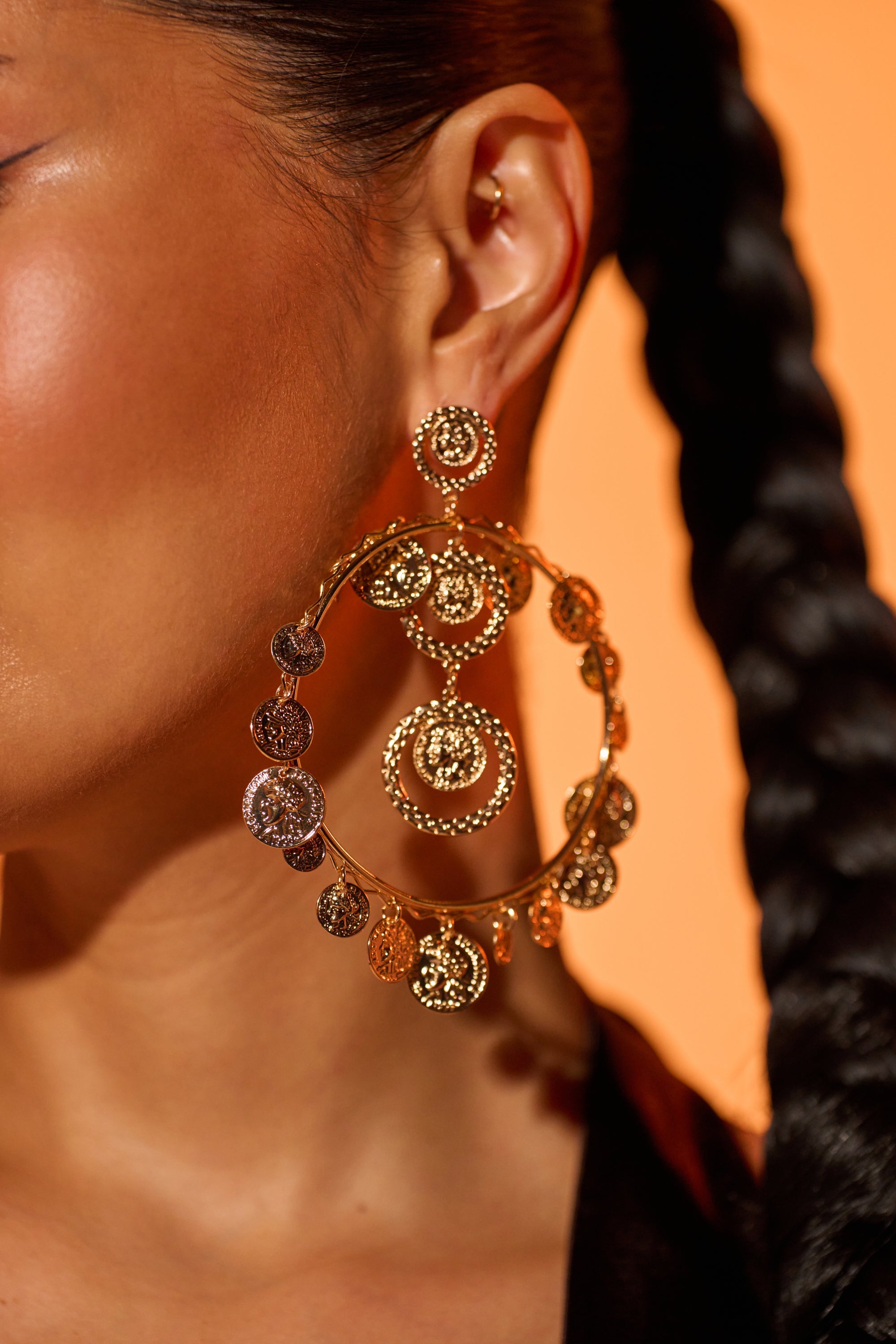 Close-up of a person's ear wearing BOHO EARRING—antique gold, large hoop earrings with multiple circular, textured designs. The background is orange, and the person has braided hair. 0