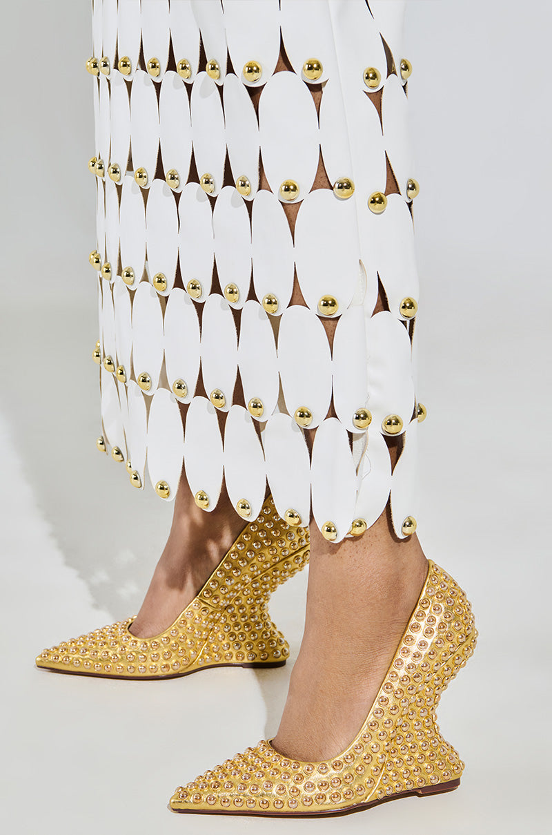 Wearing a white skirt with oval cutouts and golden studs, paired with the AZALEA WANG TERYN GOLD WEDGE PUMP in pointed toe heels, the look is contemporary and stylish, featuring bold fashion elements in metallic vegan leather.