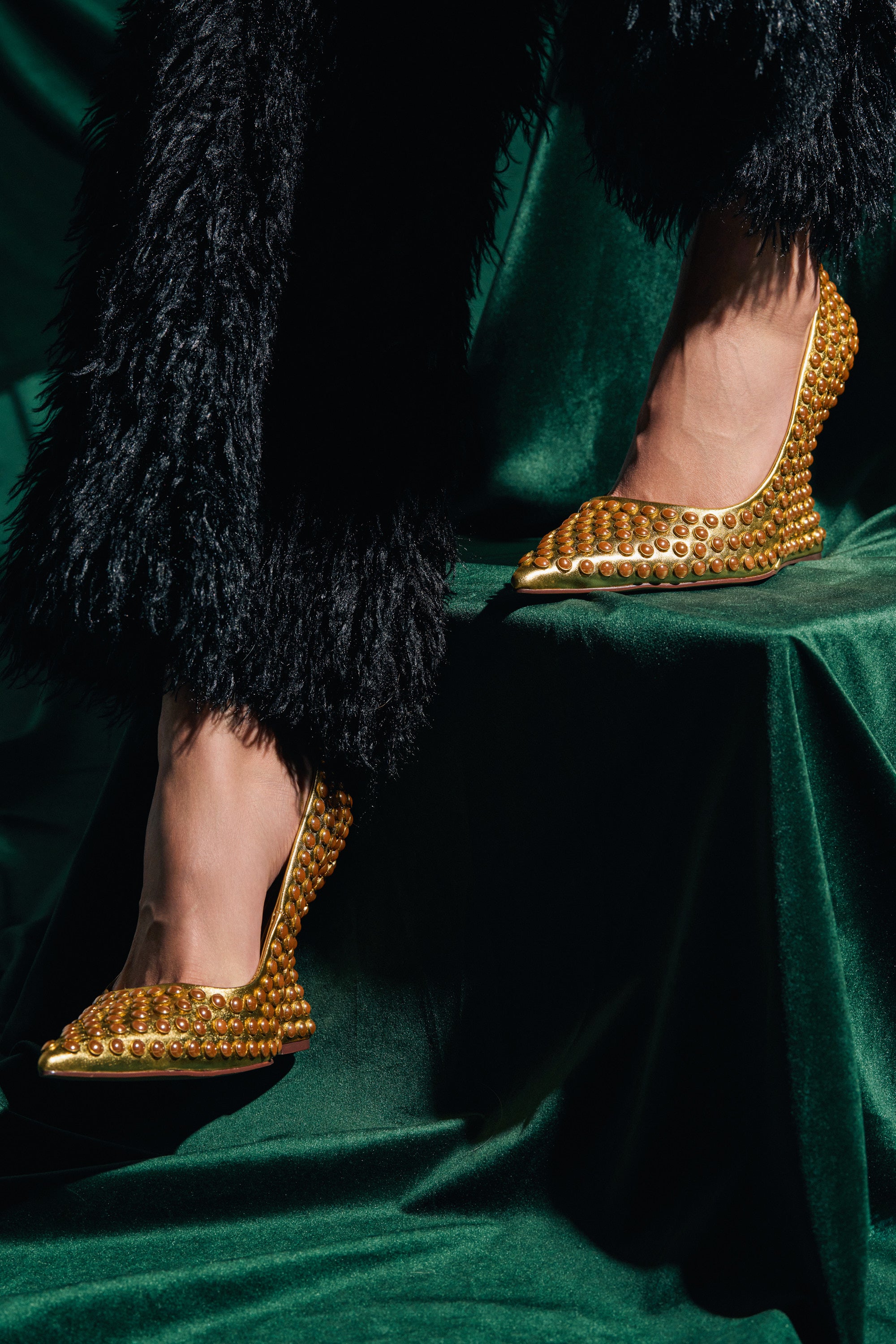 A person stands on a dark green velvet surface in textured black pants and the AZALEA WANG TERYN GOLD WEDGE PUMPS, with dramatic lighting accentuating the metallic vegan leather and rich fabric textures.