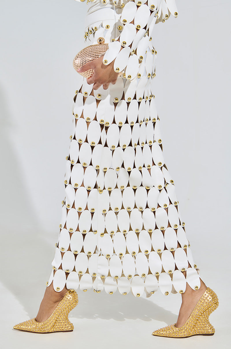 Someone wears a long white dress with oval cutouts and gold accents, paired with the AZALEA WANG TERYN Gold Wedge Pumps. They hold a small textured pink clutch in metallic vegan leather against a plain light-colored background.