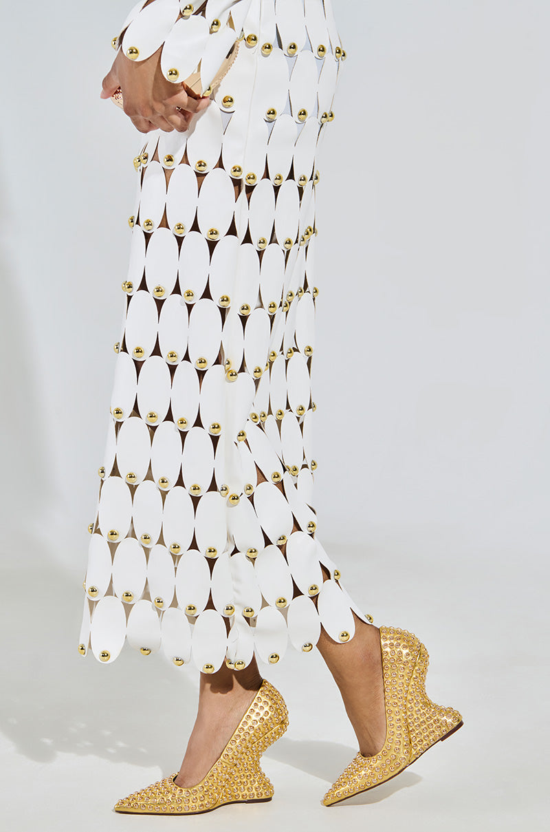 A person in a dress of white elliptical discs linked by gold rings stands with arms crossed, showcasing unique AZALEA WANG TERYN GOLD WEDGE PUMPs made from metallic vegan leather, against a plain backdrop.