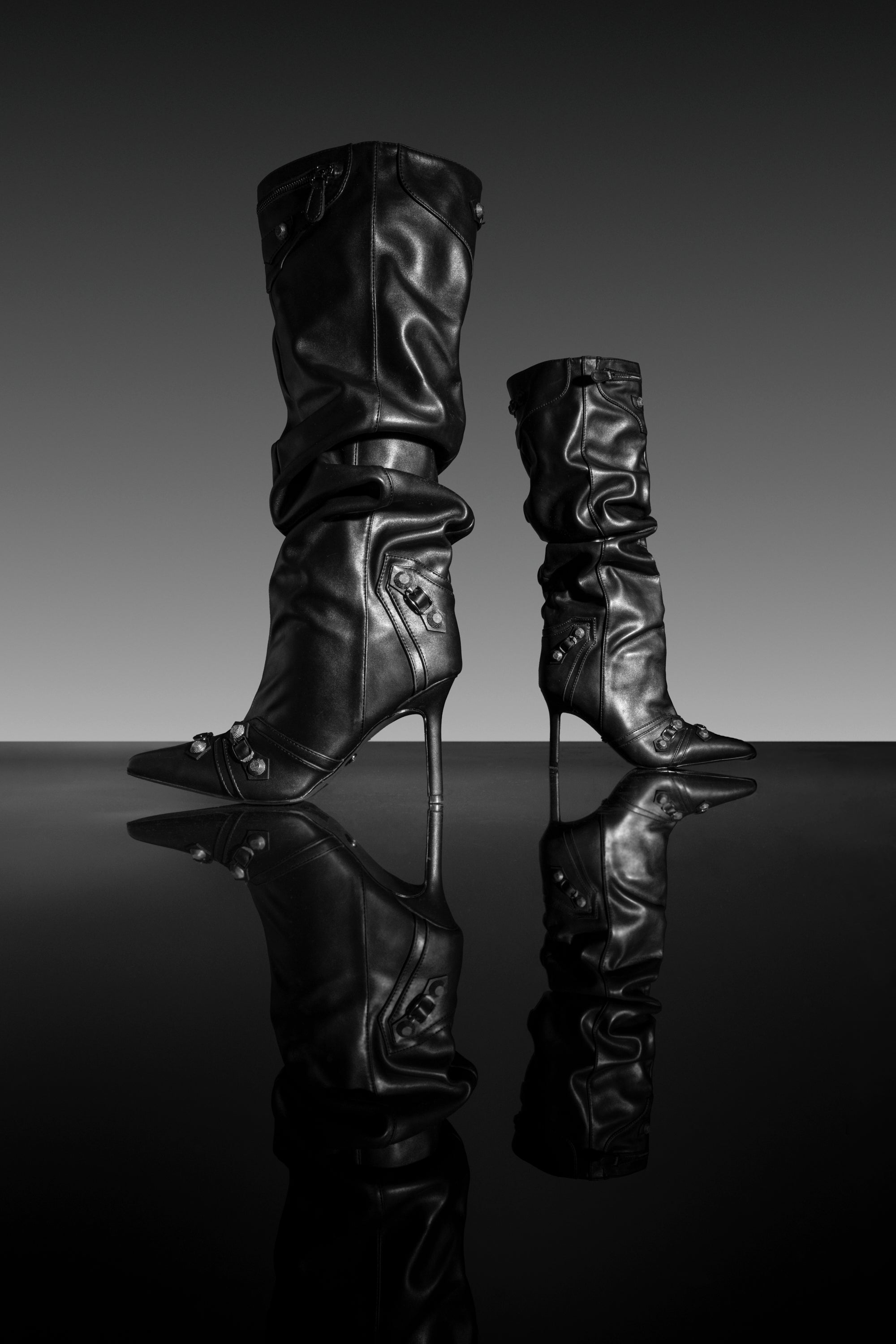 A pair of AZALEA WANG TESARA POINTED TOE HARDWARE EMBELLISHED BOOTS, featuring tall black faux leather with a slouchy stiletto silhouette, pointed toes, and stylish hardware details, stand side by side on a reflective surface against a gradient gray background.