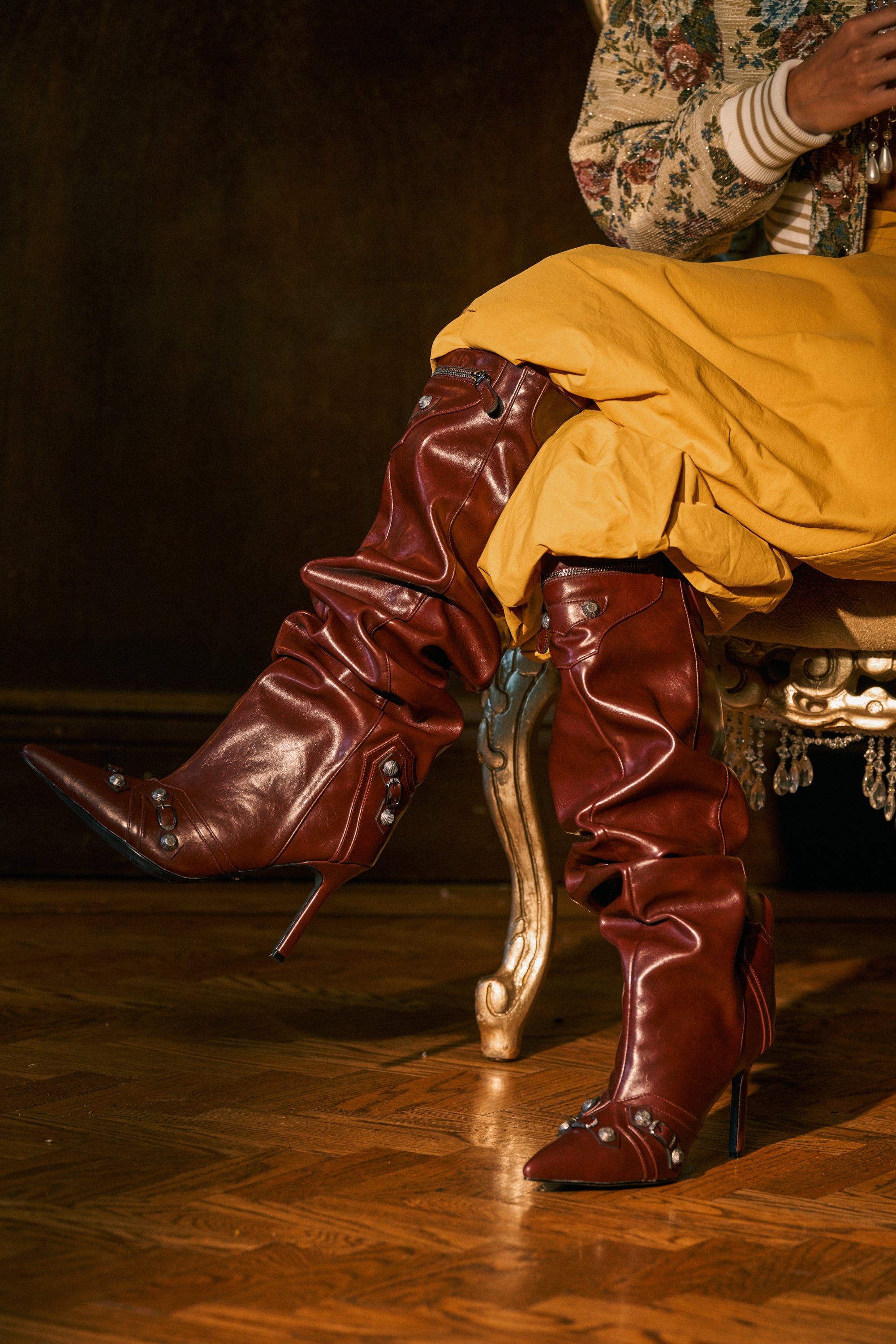 A person seated on an ornate chair, wearing yellow pants and the AZALEA WANG TESARA Burgundy Ruched Stiletto Boot with Hardware—stylish, slouchy boots adorned with decorative buttons—on a wooden floor. Upper body partially visible.