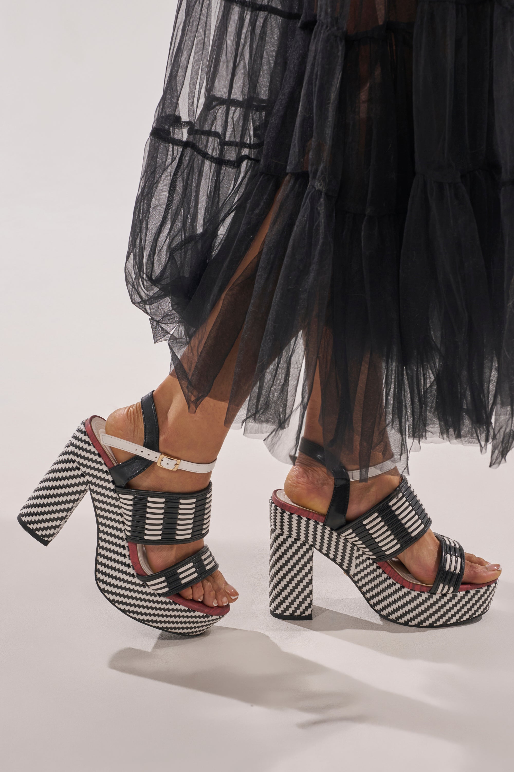 A person in a sheer black layered skirt walks confidently on a light-colored floor, wearing AZALEA WANG TESERA BLACK WHITE PLATFORM WOVEN SANDALS with bold zigzag and striped patterns and a woven platform heel. 0