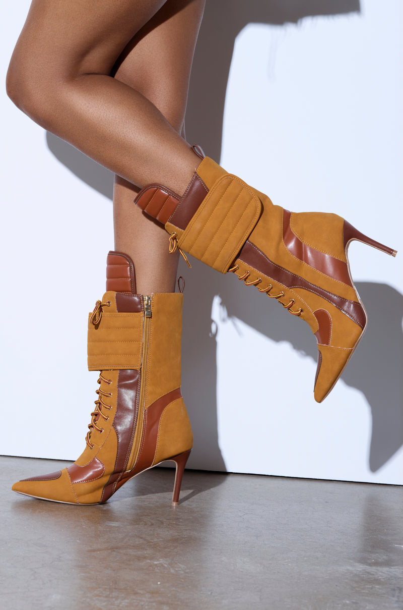 A person walks confidently in TESS CAMEL STILETTO BOOTIE high-heeled, lace-up boots with tan and brown leather. These pointed-toe boots feature side zippers and padded cuffs, set against a plain white wall and smooth concrete floor. 1