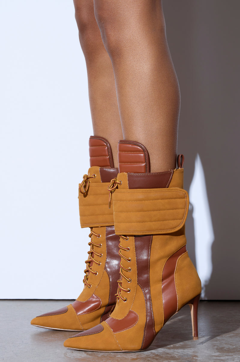 TESS CAMEL STILETTO BOOTIE: Stylish two-toned tan and brown leather high-heeled boots with lace-up fronts, quilted details, pocket-like flaps, and a pointed toe worn on a concrete floor. 2
