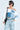 Wearing AKIRA Label sunglasses and a chic patchwork off-shoulder top, they confidently rock a black shoulder bag and distressed light-blue jeans. Their short hair highlights a bold shoulder tattoo, completing the look with an ALL AROUND THE CITY SWEATSHIRT for flair.