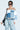 Confidently standing against a pale blue background, a person dons sunglasses and the ALL AROUND THE CITY SWEATSHIRT from the AKIRA Label. They carry a shoulder bag, wear light blue ripped jeans, and display visible tattoos on their shoulders for an edgy, modern look.