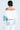 A person with short dark hair, turned away, wears the ALL AROUND THE CITY SWEATSHIRT, featuring an off-the-shoulder white cotton blend and denim sleeves. White pants with denim pockets complete the AKIRA Label ensemble, spotlighting their tattooed back against a light background.