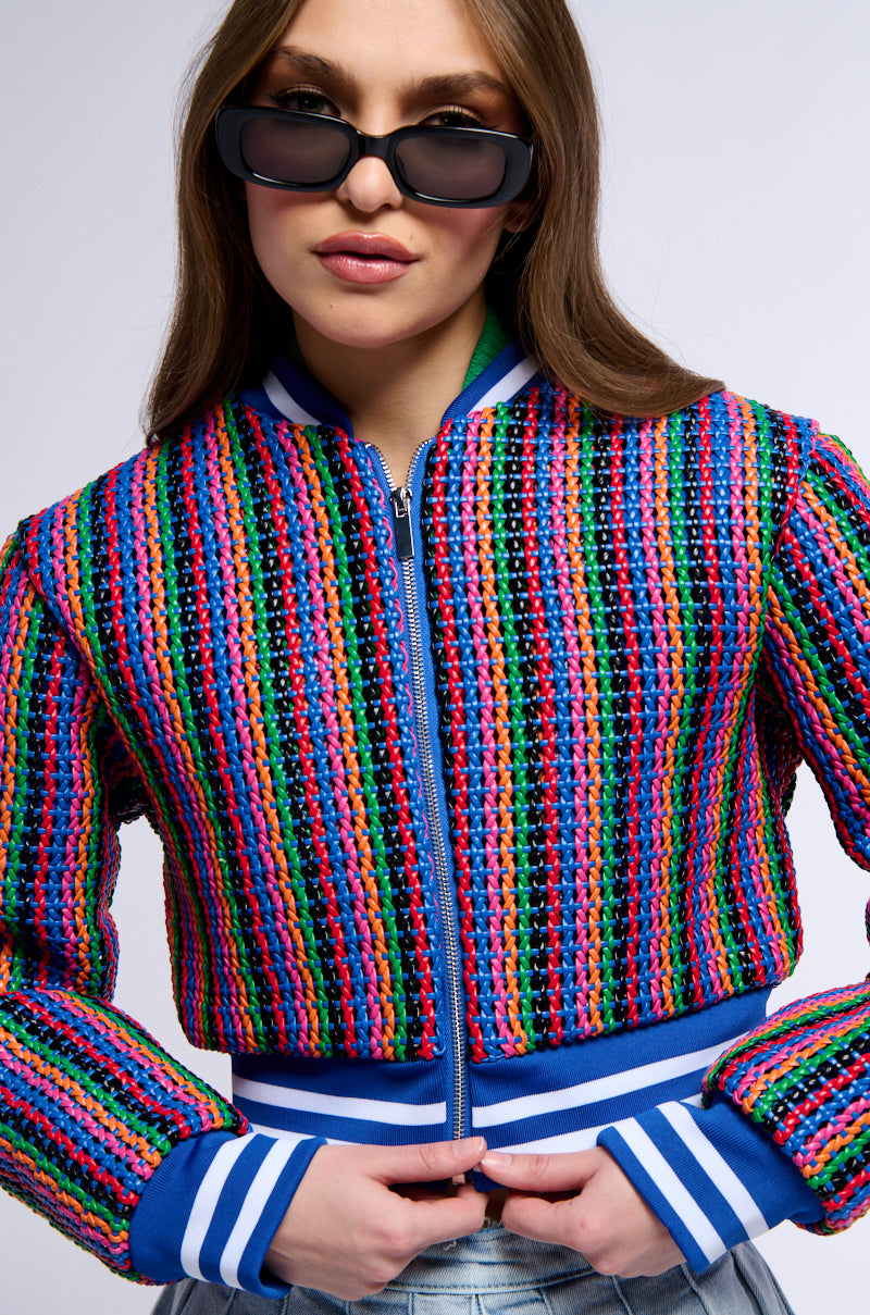 A person with long brown hair wearing black sunglasses poses confidently in the vibrant *** TEST NO GO LIVE BOMBER JACKET. This colorful, woven, striped jacket features blue and white striped cuffs, a blue zipper, and matching blue and white striped detailing at the bottom. Using SEO keywords for product visibility.