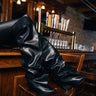 Someone wearing the AZALEA WANG TEUILA BLACK RUCHED WEDGE BOOT rests their legs on a wooden bar counter, surrounded by bar stools, beer taps, and glasses in a softly lit, lively bar atmosphere.