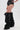 A person stands against a plain white background, showing only their lower body in black high-waisted shorts with silver studs and AZALEA WANG TEUILA BLACK RUCHED WEDGE BOOTS.