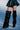 A person wearing a textured dark green skirt and AZALEA WANG TEUILA BLACK RUCHED WEDGE BOOT stands against a light blue background, with the image cropped to show only their legs and part of the skirt.
