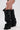 A person wears the AZALEA WANG TEUILA BLACK RUCHED WEDGE BOOT, styled with tall black ruched faux leather and a wedge heel, paired with a small black chain-strap handbag. Photo shows them from thigh down against a plain backdrop.