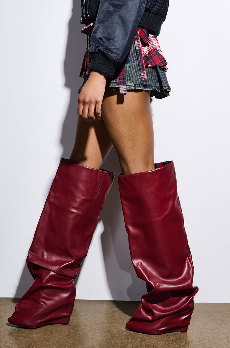 A person wears AZALEA WANG TEUILA BURGUNDY WEDGE RUCHED BOOTS reaching the thighs, paired with a short plaid skirt and black jacket. The photo focuses on the legs against a plain white wall.