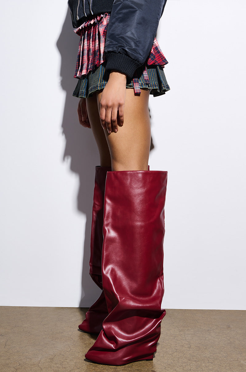 A person stands against a white wall, wearing a black top and layered red and blue plaid skirt, paired with the AZALEA WANG TEUILA burgundy wedge ruched boots that reach mid-thigh. The boots feature a slouchy faux leather design, complementing the brown floor beneath.