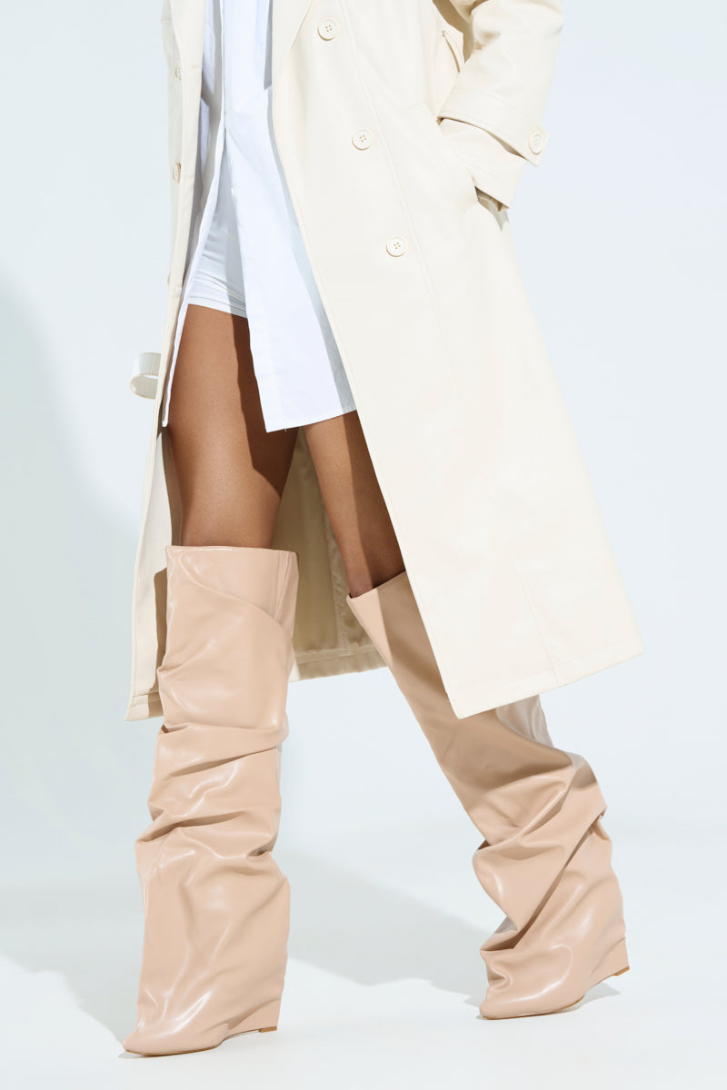 Close-up of a person wearing a long beige trench coat over a white shirt and shorts, paired with the AZALEA WANG TEUILA BONE WEDGE RUCHED BOOTS. The outfit is stylishly minimal, emphasizing the oversized boots.