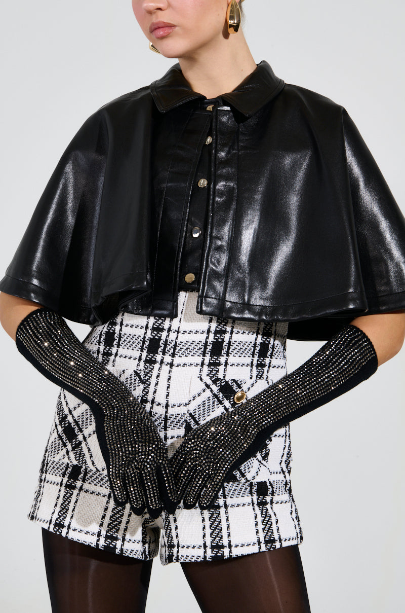 A person wearing a black leather cape, BLING BABE KNIT GLOVES IN BLACK, and black and white plaid shorts stands against a plain background. The outfit is accessorized with large hoop earrings.