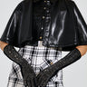 A person wearing a black leather cape, BLING BABE KNIT GLOVES IN BLACK, and black and white plaid shorts stands against a plain background. The outfit is accessorized with large hoop earrings.