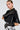 A woman with swept-back hair wears a black leather cape and the Bling Babe Knit Gloves in black, which sparkle in the light. She accessorizes with gold hoop earrings and dons a black-and-white patterned skirt, confidently posing against a plain background.