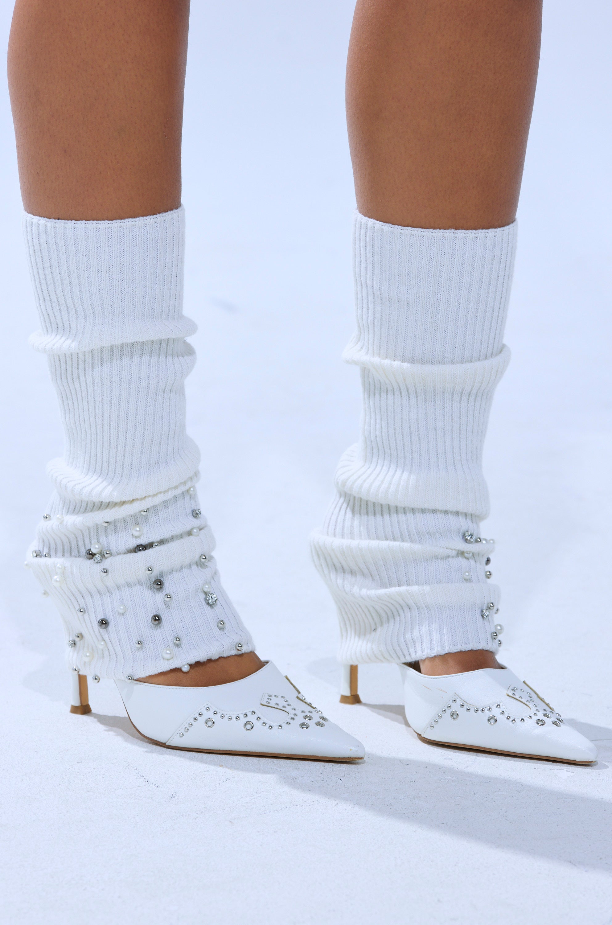 A close-up shows someone wearing COZY VIBES BLING LEGWARMERS paired with white pointed-toe heels with silver studs, standing on a light-toned surface.