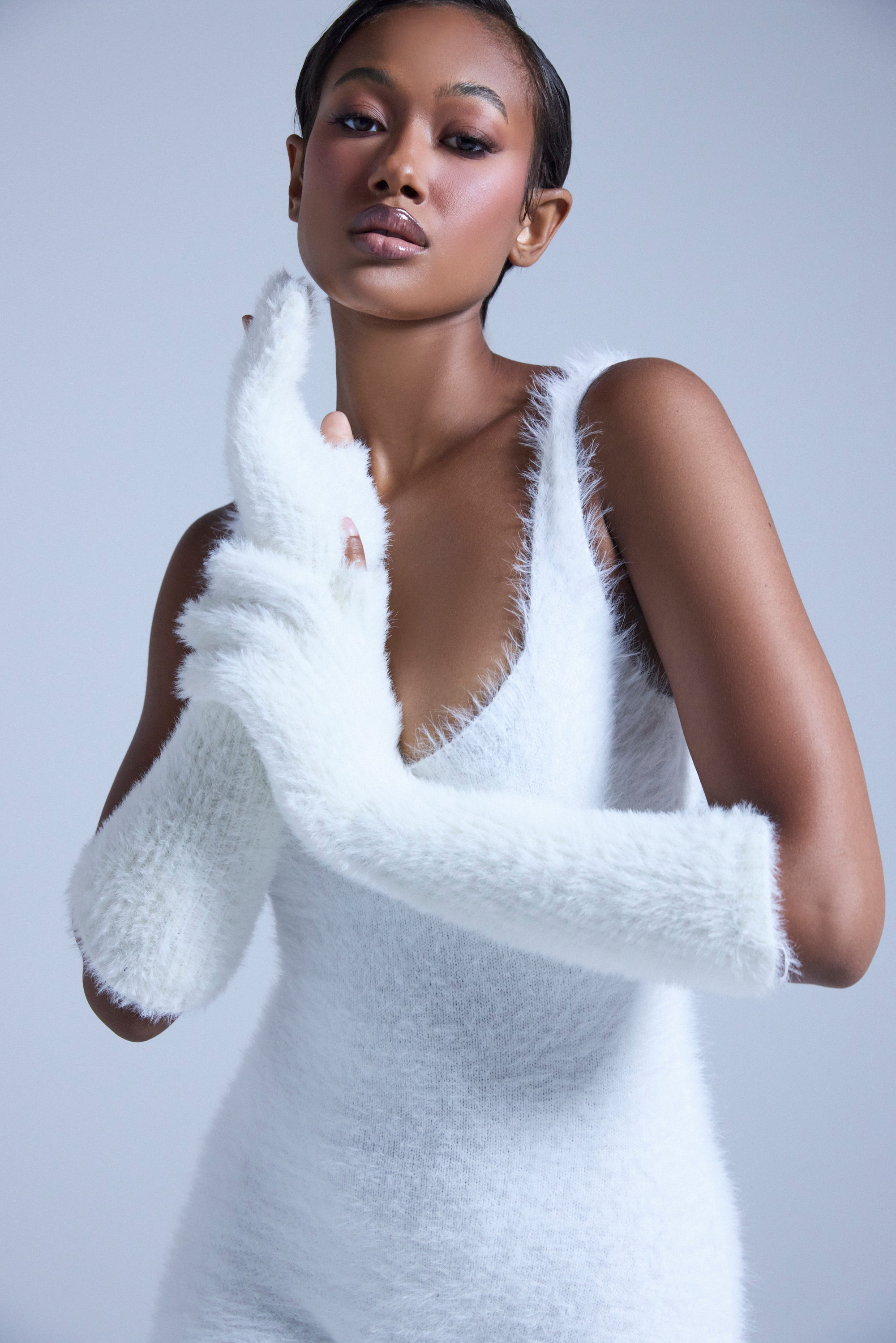 A woman poses confidently against a light background, wearing PEARLY WHITES FUZZY GLOVES with a matching fuzzy, sleeveless white dress. Her slicked-back hair and calm gaze complete the chic, modern look.