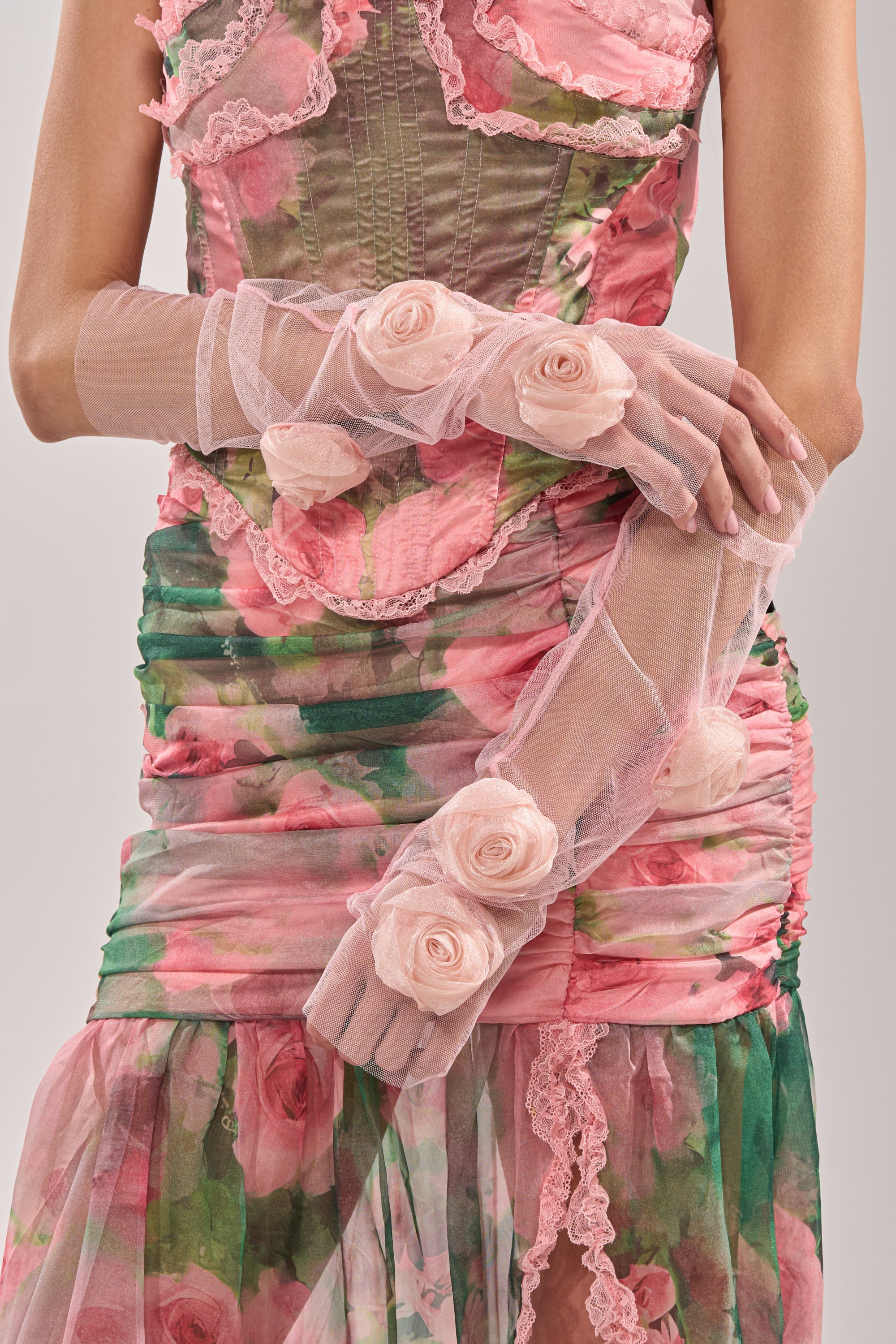 Wearing a pink and green floral dress with ruffles, lace trim, and a fitted bodice, this look is finished with SPRING SZN APPLIQUE GLOVES—sheer mesh gloves embellished with fabric roses—as the person crosses their arms at the waist. 3