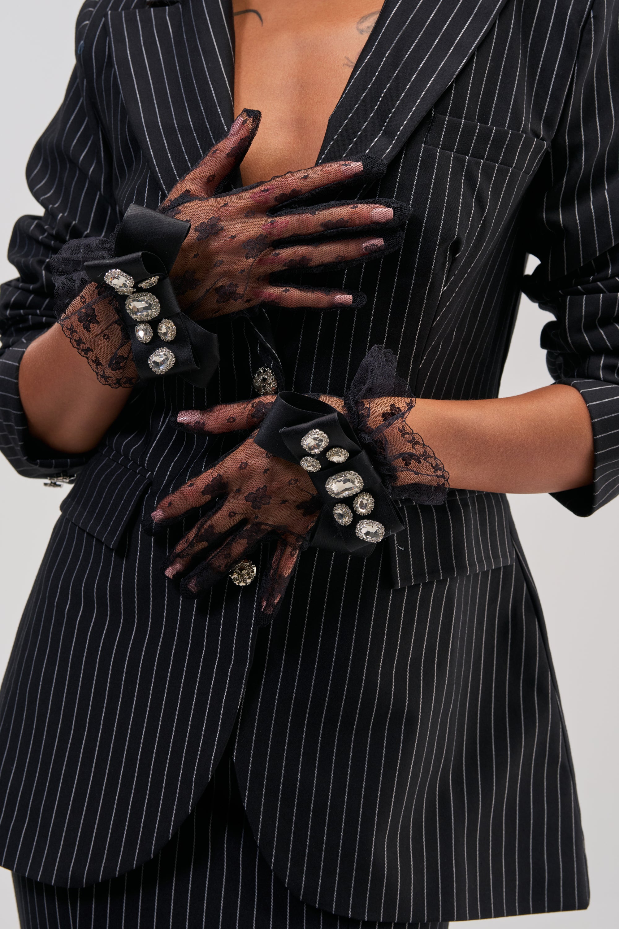 Wearing the VERY DEMURE LACE GLOVES, a person dressed in a black pinstripe suit crosses their hands over their chest and waist, showcasing the vintage-inspired black lace gloves embellished with large rhinestones against a plain background.