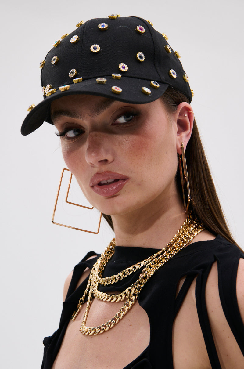A woman wearing the BLING HAT with gemstone embellishments, large square hoop earrings, layered gold chains, and a black cut-out shoulder top, styled with natural makeup, looks slightly to the side.