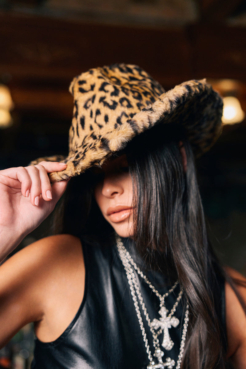 A woman with long dark hair in a black sleeveless top partially covers her face with the CHECK ME OUT FAUX FUR COWBOY HAT, a bold leopard print western accessory, styled with layered silver cross necklaces for added edge.