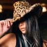 A woman with long dark hair in a black sleeveless top partially covers her face with the CHECK ME OUT FAUX FUR COWBOY HAT, a bold leopard print western accessory, styled with layered silver cross necklaces for added edge.